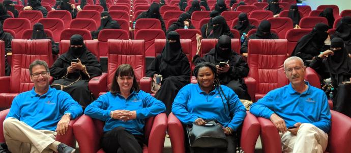 Kathy, Gary, Connie, and Ivan in an auditorium in blue shirts in front of people dressed in burkas