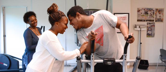 Two people in foreground, one on right is on a treadmill, they are both smiling and have heads almost touching. Another person in background is looking at them smiling 