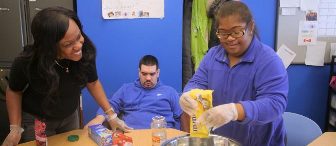 three people at a table, 2 are stood, the one on the left is looking on the person on the right who is putting candy in a bowl. The 3rd person sits in the background between them.