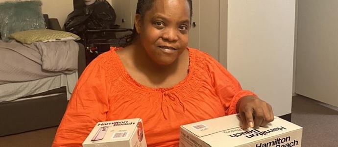 Latasha wears orange while sitting at a chair in a bedroom, she is holding boxes for small kitchen appliances