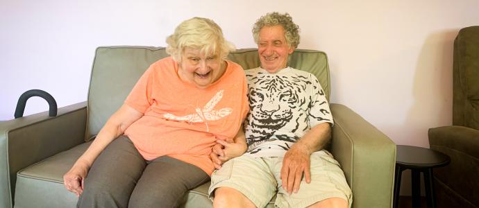 Joan and Melvin sat together on the couch, Joan is leaning forward and smiling, Melvin is looking at Joan