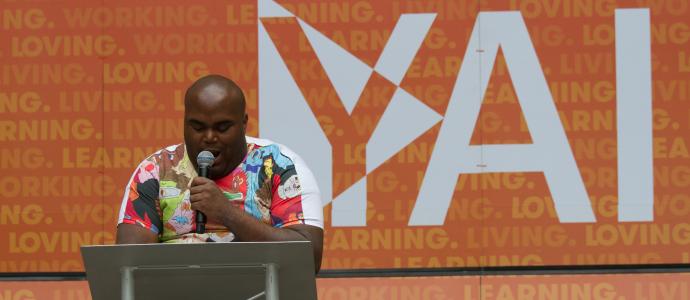 Person stands at a podium, holding microphone in front of a large YAI sign with orange background