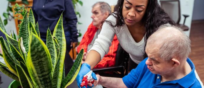 Two people work together to care for a plant, one is sat, the other is stood behind them supporting them