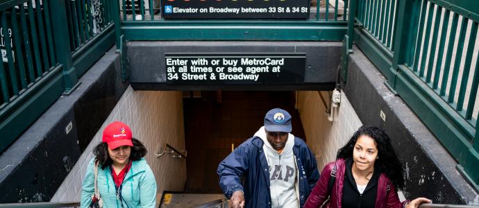 Group of people walking up the subway steps, you can see the sign for Herald Sq Station above them