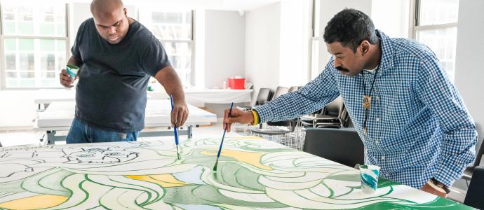 2 people painting a large mural for YAI's HQ on a tabletop.