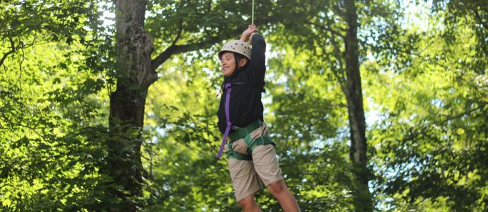 Child wearing shorts, long sleeved top and helmet is on a zipline in the trees.