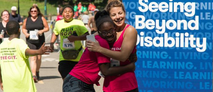 2 people hug at the finish line of 5k run at central park challenge. You see a sign on the right that says "seeing beyond disability"