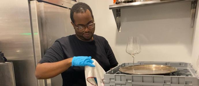 Jawann is standing in front of a fridge, drying a wine glass and a tray using a towel.