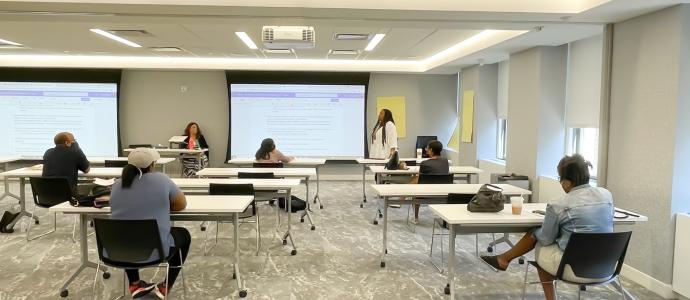 Training room with people safely distanced for COVID. Consuela Senior stands at the front of the room and you see 2 projected training documents at the front of the room too.