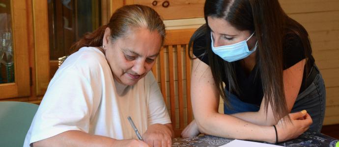 Two people sit at at table. One on the left is writing on a piece of paper, the other on the right is looking on and wears a mask.