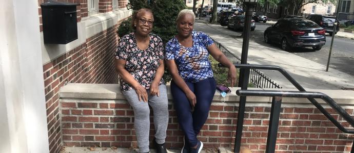 2 people sit on a wall outside an apartment building, they are posing for the photo