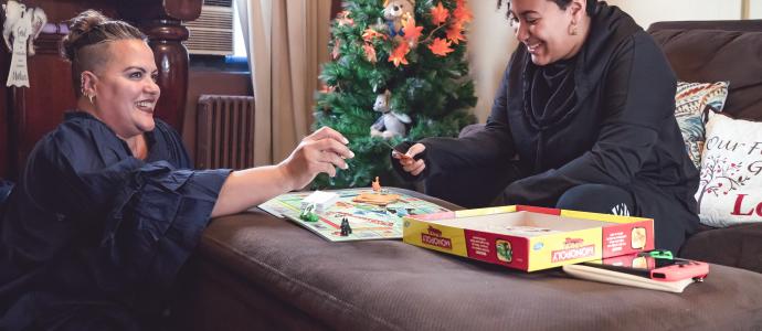 Two smiling people in a home playing monopoly. One sits on the floor the other is on a couch