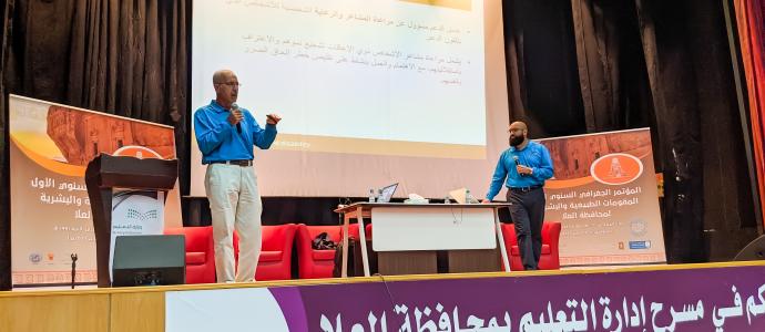 Two YAI Training Staff stand on a stage in front of a powerpoint, all text on the powerpoint is in Arabic