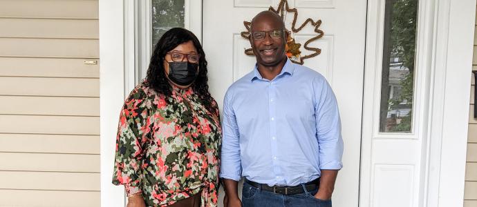 Two people stand in front of a door, one is wearing a flowery shirt and mask, the other is wearing a light blue button down shirt.