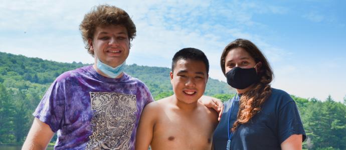Two kids and one camp counselor (wearing a mask) stand together side by side and post for photo. You see green trees behind them