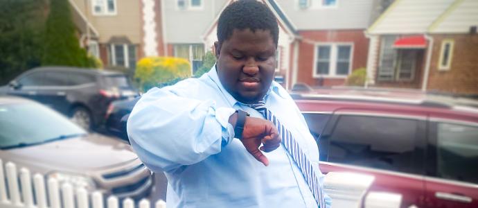 Person in blue button down shirt and tie stands outside looking at his watch. You see houses and cars in the background