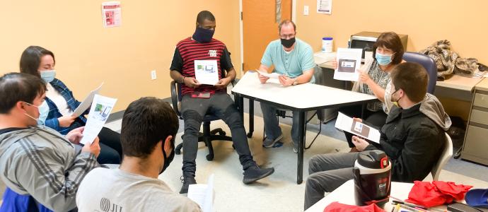 Group of people sitting in a circle in a room, they are holding paper or brochures in their hands and wearing masks