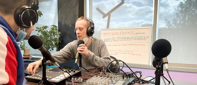 Two people sit at a table with mics and a mixing desk, they are wearing headphones and appear to be talking