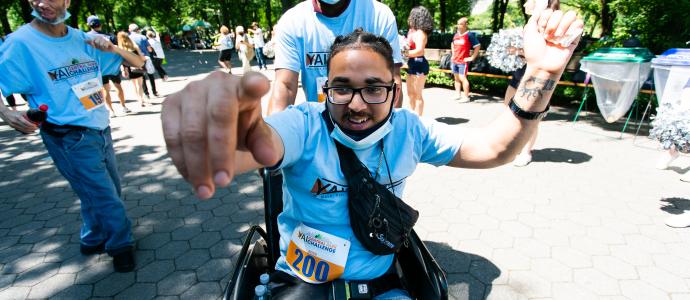 Young person in a wheelchair points at camera with left arm up. Someone else stands behind them. There are people all around in in the background in Central Park