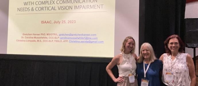 3 women stand on a stage in front of a projected image of presentation