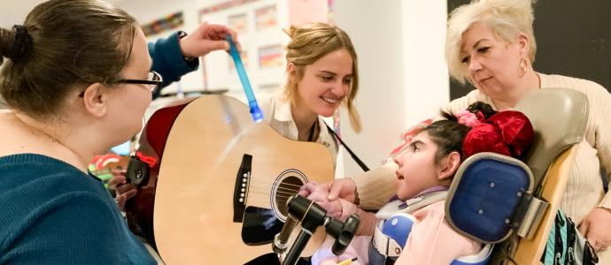 Staff gather around a student in a wheelchair, they are playing with a guitar