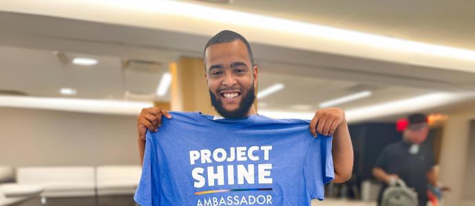 Breon smiles and poses for photo holding up a project SHINE t-shirt