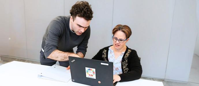 Two people. one stood next to the other who is sat at a desk, they are both looking at and interacting with a laptop