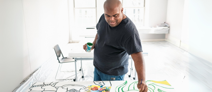 Jimmy stands in a relatively empty room with a large canvas in front of him. He holds a cup of paint in one hand and a paintbrush in the other