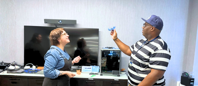 Two people stand in a room with various technological items. They are facing one another while the person on the right holds up a 3D printed item