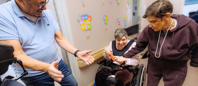 3 people at a day program, they look happy and engaged as 2 look on at the person in the middle who is in a wheelchair.