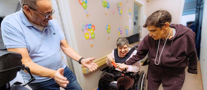 3 people at a day program, they look happy and engaged as 2 look on at the person in the middle who is in a wheelchair.