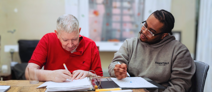 Two men sit at a table, they are both holding pens and have paper in front of them like they are drawing or writing