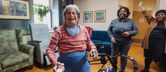 woman stands by her walker mid laughter. behind her to the right are 2 staff members laughing and celebrating her