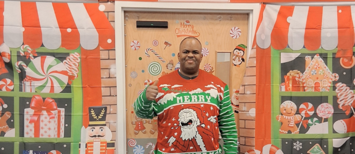 Kenneth stands and poses in his ugly Christmas sweater in front of a Christmas-themed backdrop