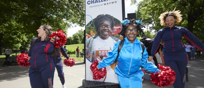 A person supported by YAI is holding red pom poms and dancing with NY Cheer cheerleaders in front of a CPC sign with their picture.
