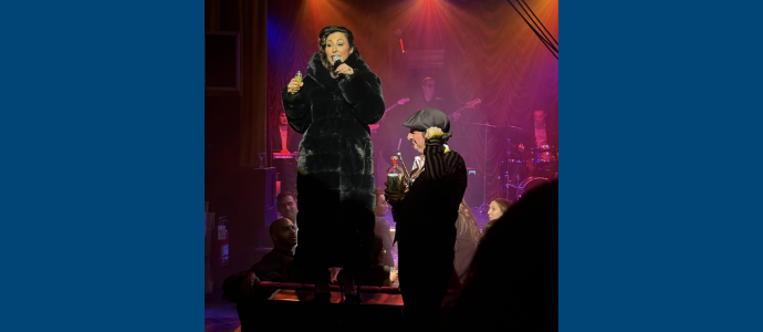 Angie Pontani in a dark fur coat sings into a microphone on a stage while another performer stands front; band members and drum equipment sit behind them under colorful spotlights.