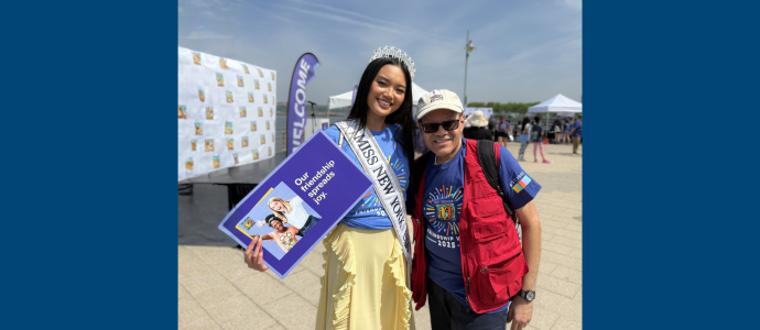 Two people smiling outdoors at an event, Miss New York is wearing a sash and a crown-like headpiece and holding a program booklet, standing beside Ismael in a cap and red vest on a sunny day.