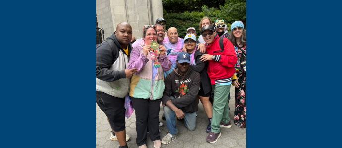 Group of Self Advocates posing outdoors by the bandshell in Central Park and wearing colorful clothing, with one person holding a small medal award.