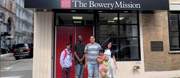 Group of people standing in front of a storefront entrance labeled “The Bowery Mission” with glass doors and brick walls on a New York City street.