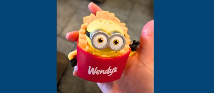 Ashley's hand holds a small red and yellow “Wendy’s” minion toy (minion in fries with large googly-style glasses)