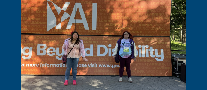 Ashley (right) and her sister stand in central park in front of a large orange sign with “YAI Seeing Beyond Disability,” text.