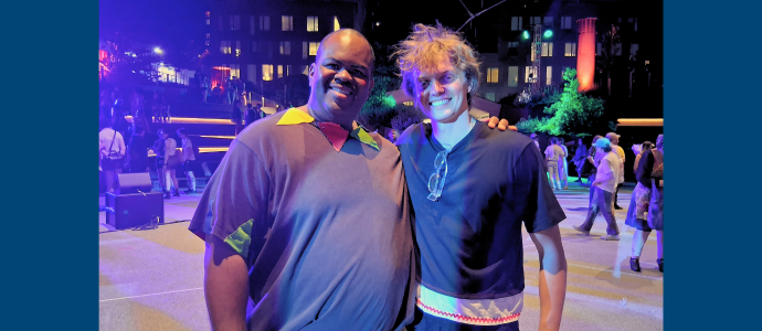 Kenneth stands with Tim, they are smiling and standing close together at night on a city street, with colorful event lighting and tall apartment buildings in the background.