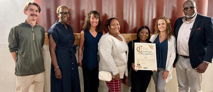 Group of seven adults posing indoors, one holding a framed certificate of recognition.