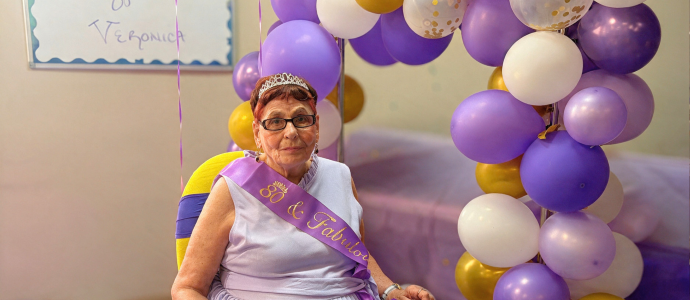 Veronica is seated indoors at her 80th birthday celebration. She is wearing a purple sash and glasses, with a large purple, white, and gold balloon cluster behind her and a hand-drawn “Happy Birthday” message on the wall.