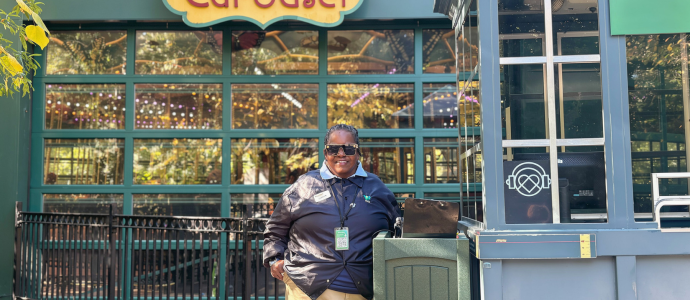 Person standing near a gray ticket booth in front of a colorful “Bug Carousel” sign, with a large glass building behind and autumn leaves framing the scene.