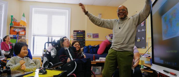 Man stands with arms in air in front of a smartboard and children and staff in classroom