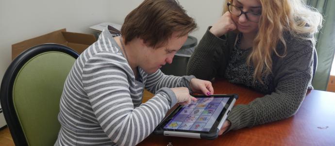 two women sit at a table, one is using an ipad while the other watches/supports