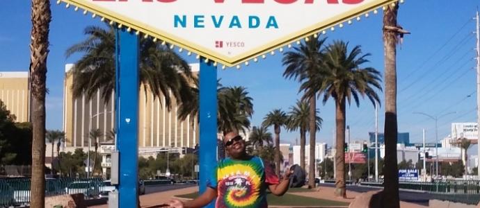 Omari wears a tie-dye shirt and stands in front of the welcome to Las Vegas sign