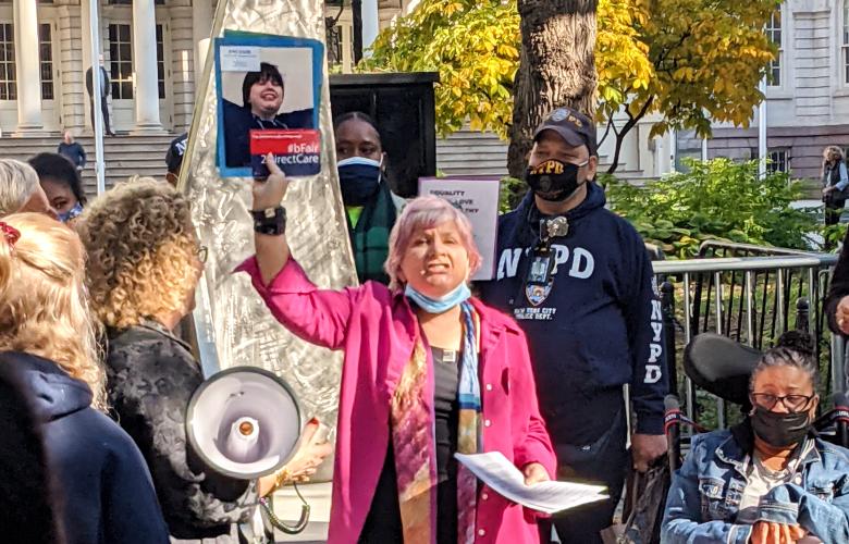 Woman stands in the middle of a group of people to appear to be lobbying/protesting, she holds up a photograph of someone, another person is holding a megaphone, someone on her left is in a wheelchair. There's an NYPD officer stood in the background