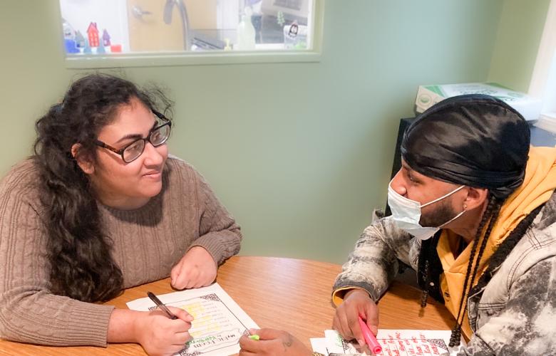 Two people sit at a desk, Karima on the left is writing, they are both looking at each other